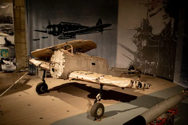 Photograph of WWII aircraft Nakajima B5N “Kate”. Featured sitting on platform inside of exhibit with photo backdrop.
