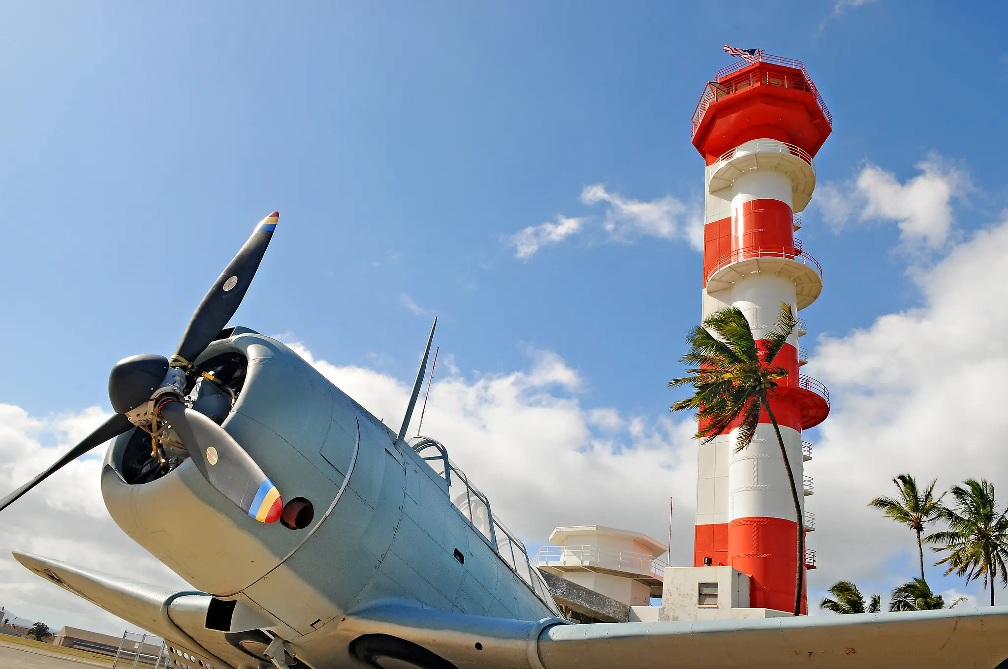 Photograph of the front of an aircraft in the foreground and the Ford Island Control Tower standing proudly in the background. Taken after recent rennovation.