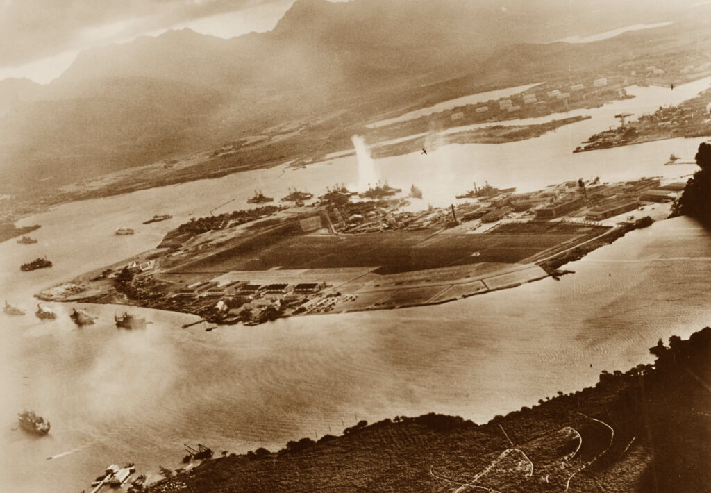 Photograph taken from a Japanese plane during the torpedo attack on ships moored on both sides of Ford Island. View looks about east, with the supply depot, submarine base and fuel tank farm in the right center distance. A torpedo has just hit USS West Virginia on the far side of Ford Island (center). Other battleships moored nearby are (from left): Nevada, Arizona, Tennessee (inboard of West Virginia), Oklahoma (torpedoed and listing) alongside Maryland, and California. On the near side of Ford Island, to the left, are light cruisers Detroit and Raleigh, target and training ship Utah and seaplane tender Tangier. Raleigh and Utah have been torpedoed, and Utah is listing sharply to port. Japanese planes are visible in the right center (over Ford Island) and over the Navy Yard at right. Japanese writing in the lower right states that the photograph was reproduced by authorization of the Navy Ministry. U.S. Naval History and Heritage Command Photograph.