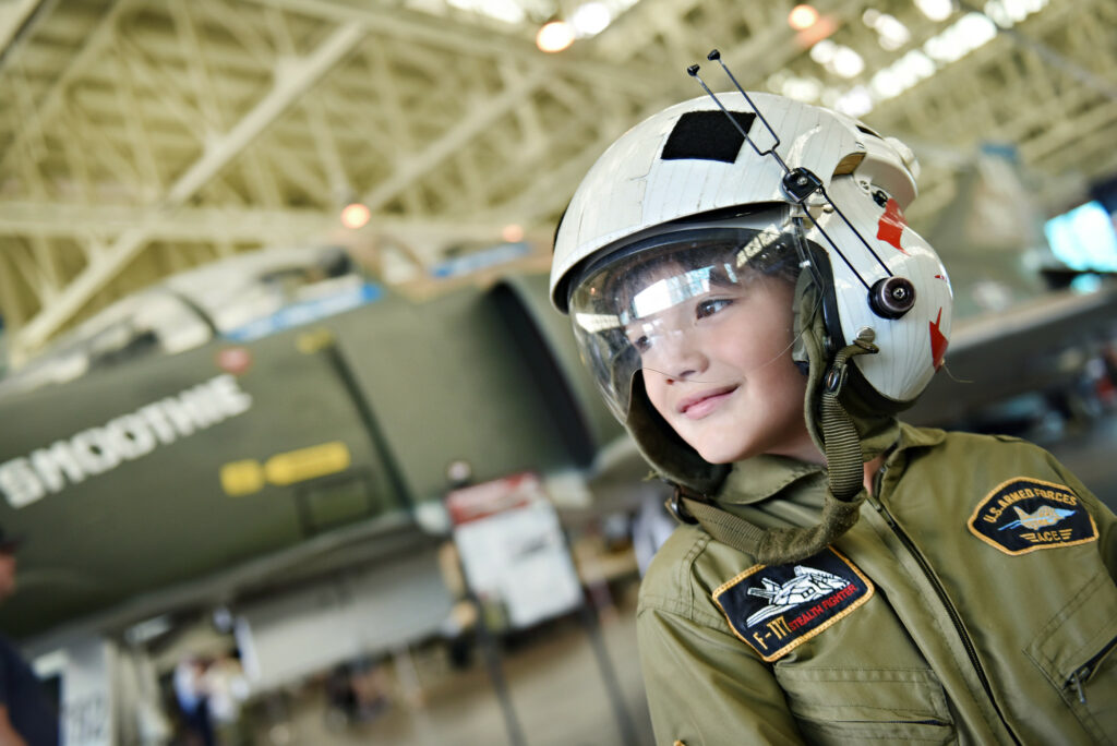 Photo of child dressed up with a helmet standing in front of a plane