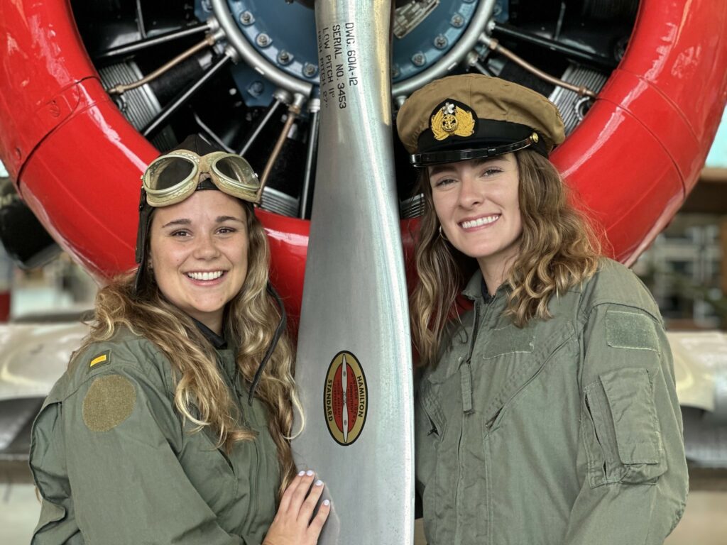 Two women in pilot costumes smiling while posing near the rotor of an aircraft.